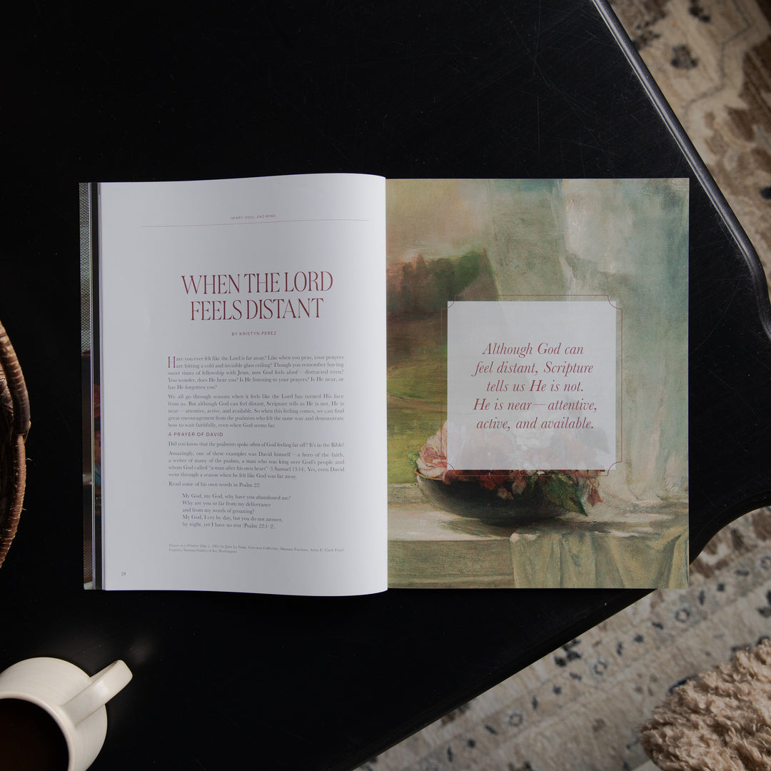 An open book or magazine laid on a dark tabletop, showing a page titled 'WHEN THE LORD FEELS DISTANT' on the left and a scripture art page on the right. A white coffee cup is partially visible at the bottom left, and patterned rugs appear around the table.