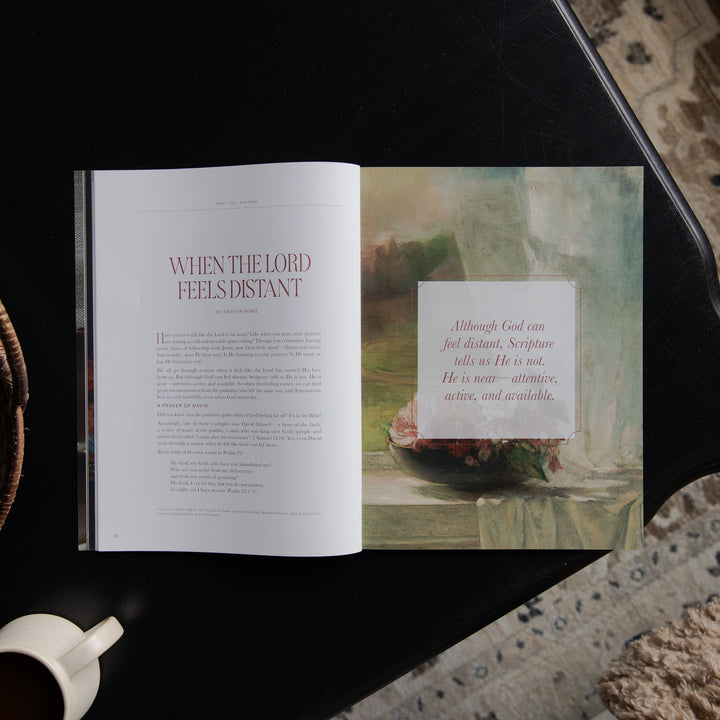 An open book or magazine laid on a dark tabletop, showing a page titled 'WHEN THE LORD FEELS DISTANT' on the left and a scripture art page on the right. A white coffee cup is partially visible at the bottom left, and patterned rugs appear around the table.
