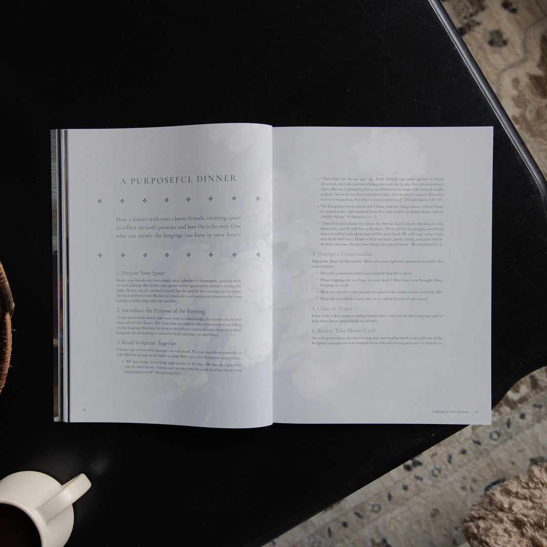 An open book or magazine spread laid on a dark tabletop, showing two white pages with printed text and decorative dotted line details. A partial white cup is visible at the lower left, and a textured stone or concrete surface appears at the right edge.