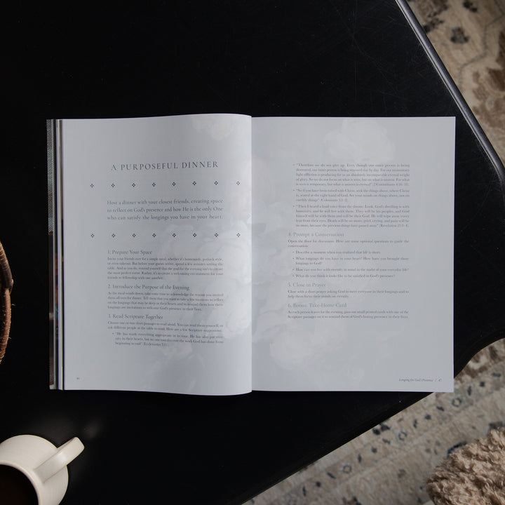 An open book or magazine spread laid on a dark tabletop, showing two white pages with printed text and decorative dotted line details. A partial white cup is visible at the lower left, and a textured stone or concrete surface appears at the right edge.