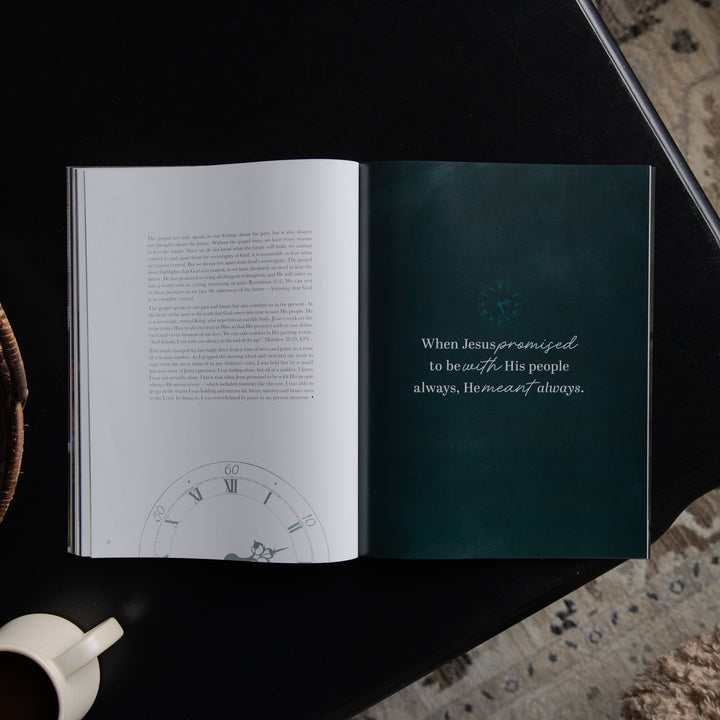 An open book laid flat on a dark surface, showing a white left page with dense text and a clock illustration near the bottom, and a dark teal right page with a quote in white script. A white mug is partially visible at the lower left, and a patterned rug appears at the lower right.
