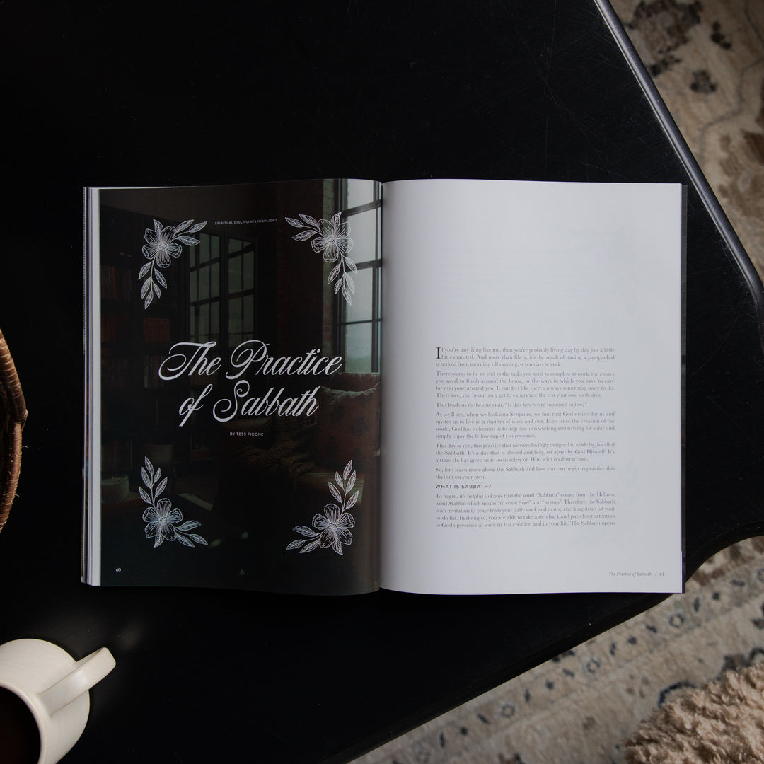 An open book titled "The Practice of Sabbath" lies on a dark tabletop, with the left page showing a black cover decorated with white floral illustrations and the right page displaying printed text. A white cup is partially visible at the lower left, and a patterned rug appears at the lower right.