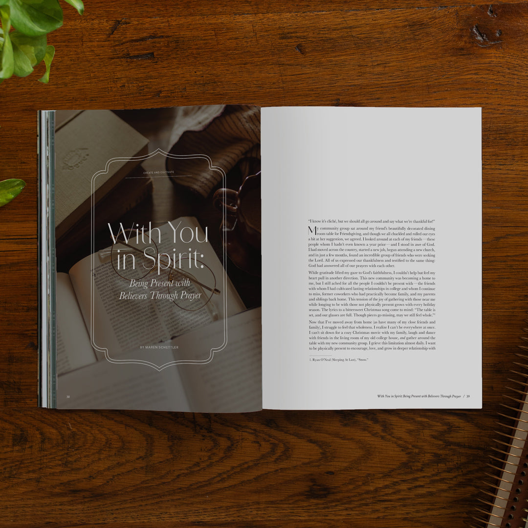An open printed magazine spread laid flat on a dark wooden tabletop. The left page shows a warm-toned photo of an open book with the title 'With You in Spirit'.