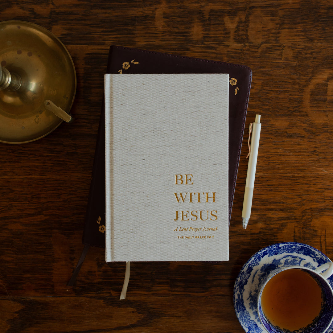 Be With Jesus Lent Prayer Journal cover page on a wooden table and on a closed bible next to a bronze candle lamp, a tea cup, and a pen