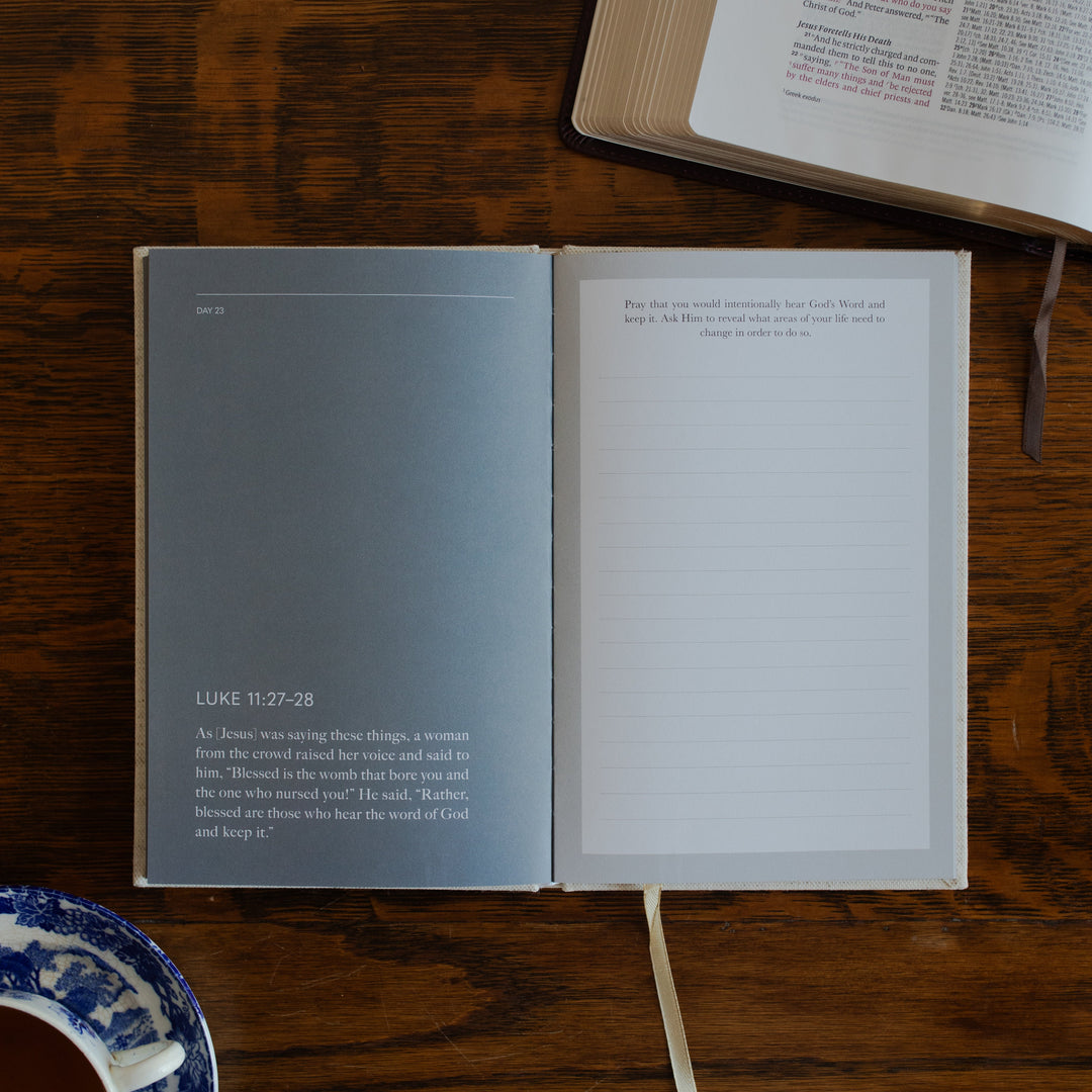 Be With Jesus Lent Prayer Journal opened to the Luke 11:27 questions page on a wooden table next to an open bible and a tea cup