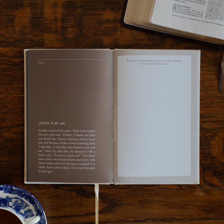 Be With Jesus Lent Prayer Journal opened to the John 11:41 questions page on a wooden table next to an open bible and a tea cup