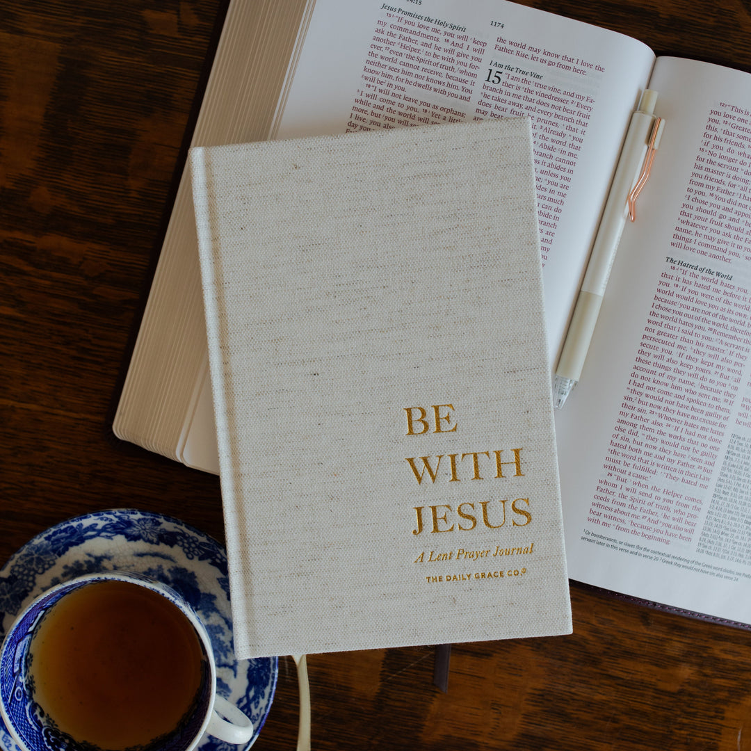 Be With Jesus Lent Prayer Journal cover page on a wooden table and on a open bible next to a tea cup