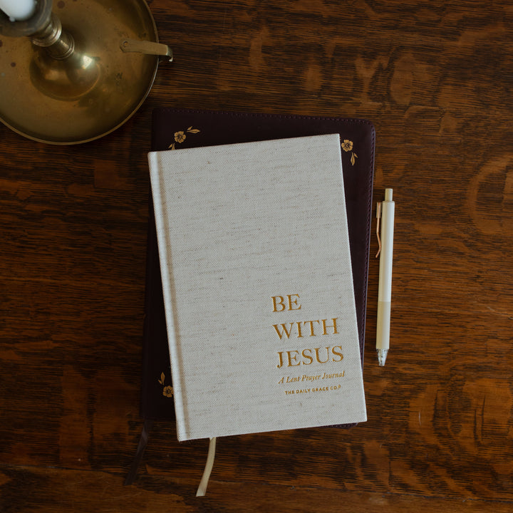 Be With Jesus Lent Prayer Journal cover page on a wooden table and on a closed bible next to a bronze candle lamp