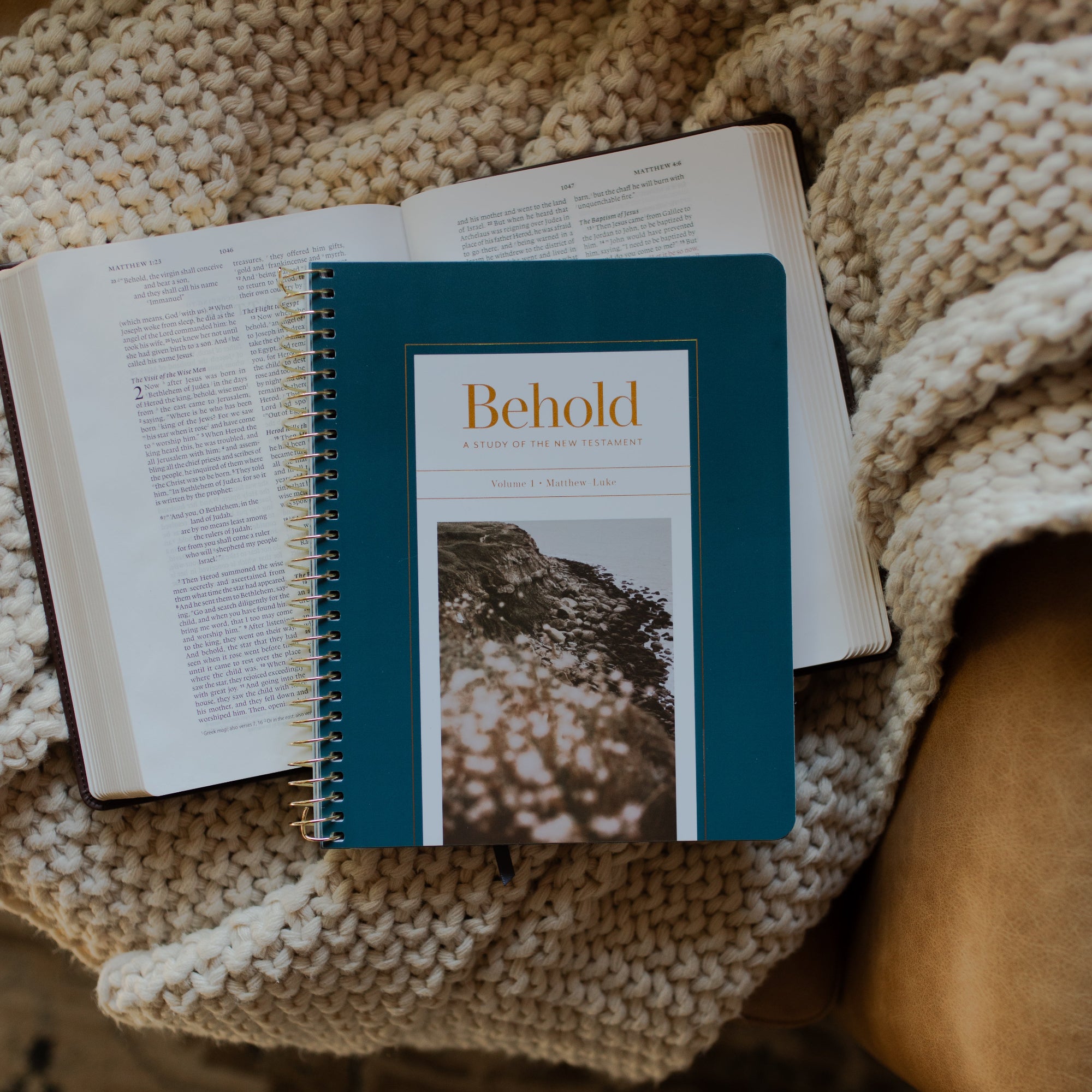 Behold Volume 1 Spiral Study cover on an open bible on a knitted blanket