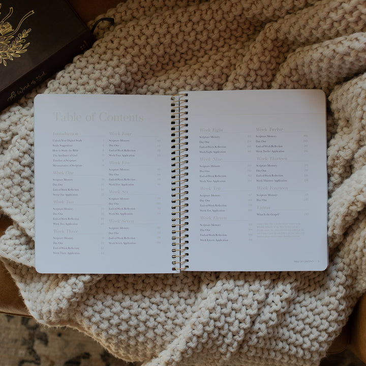 Behold Volume 1 Spiral Study opened to the table of contents page on a knitted blanket next to a bible