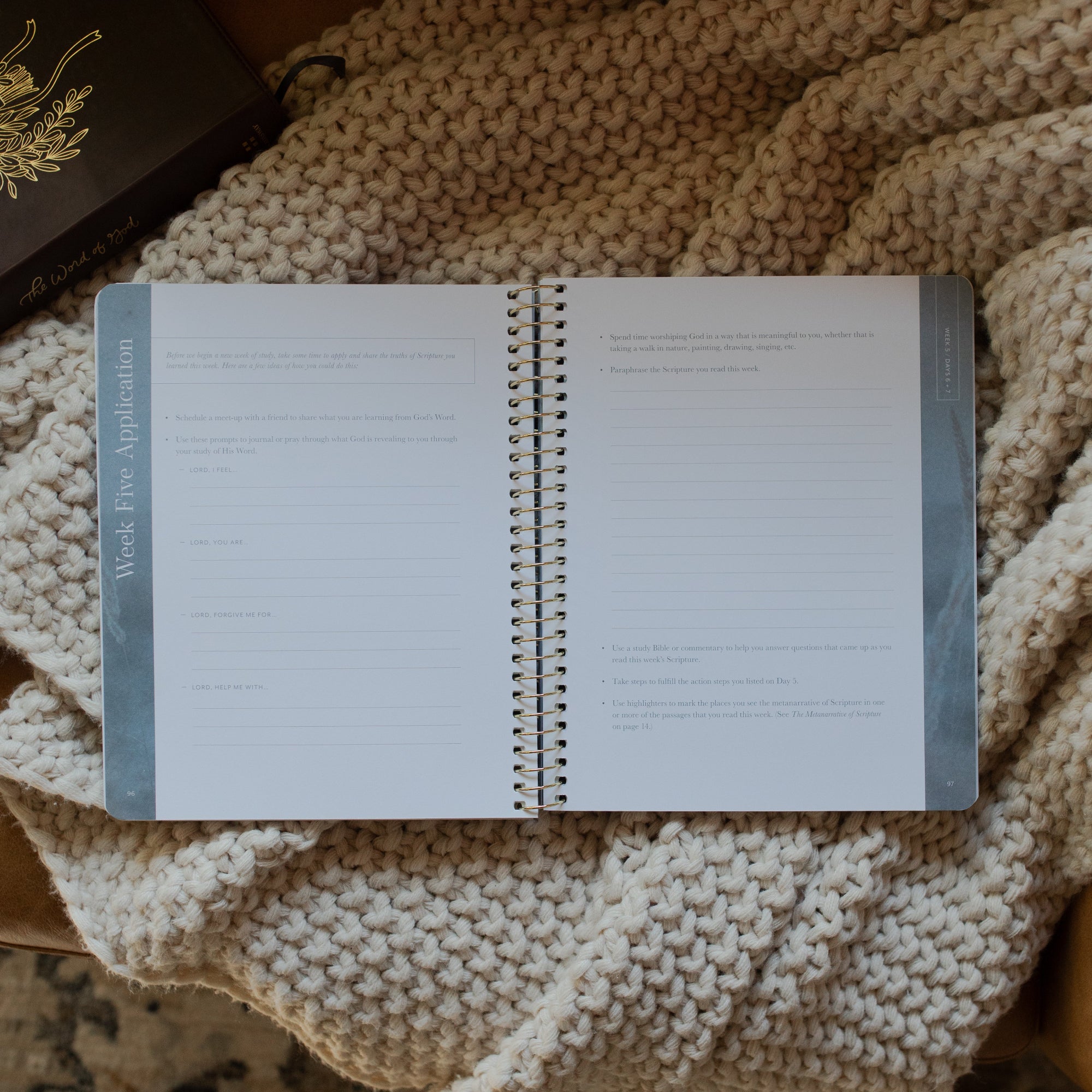 Behold Volume 1 Spiral Study opened to the week 5 applications page on a knitted blanket next to a bible