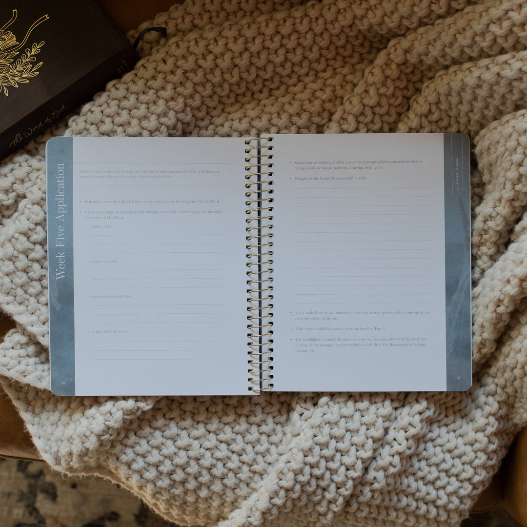 Behold Volume 1 Spiral Study opened to the week 5 applications page on a knitted blanket next to a bible