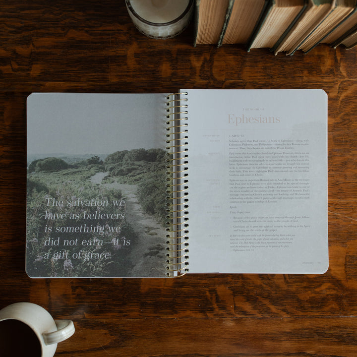 Behold Spiral opened to the book of Ephesians page on a wooden table next to a coffee cup and some books