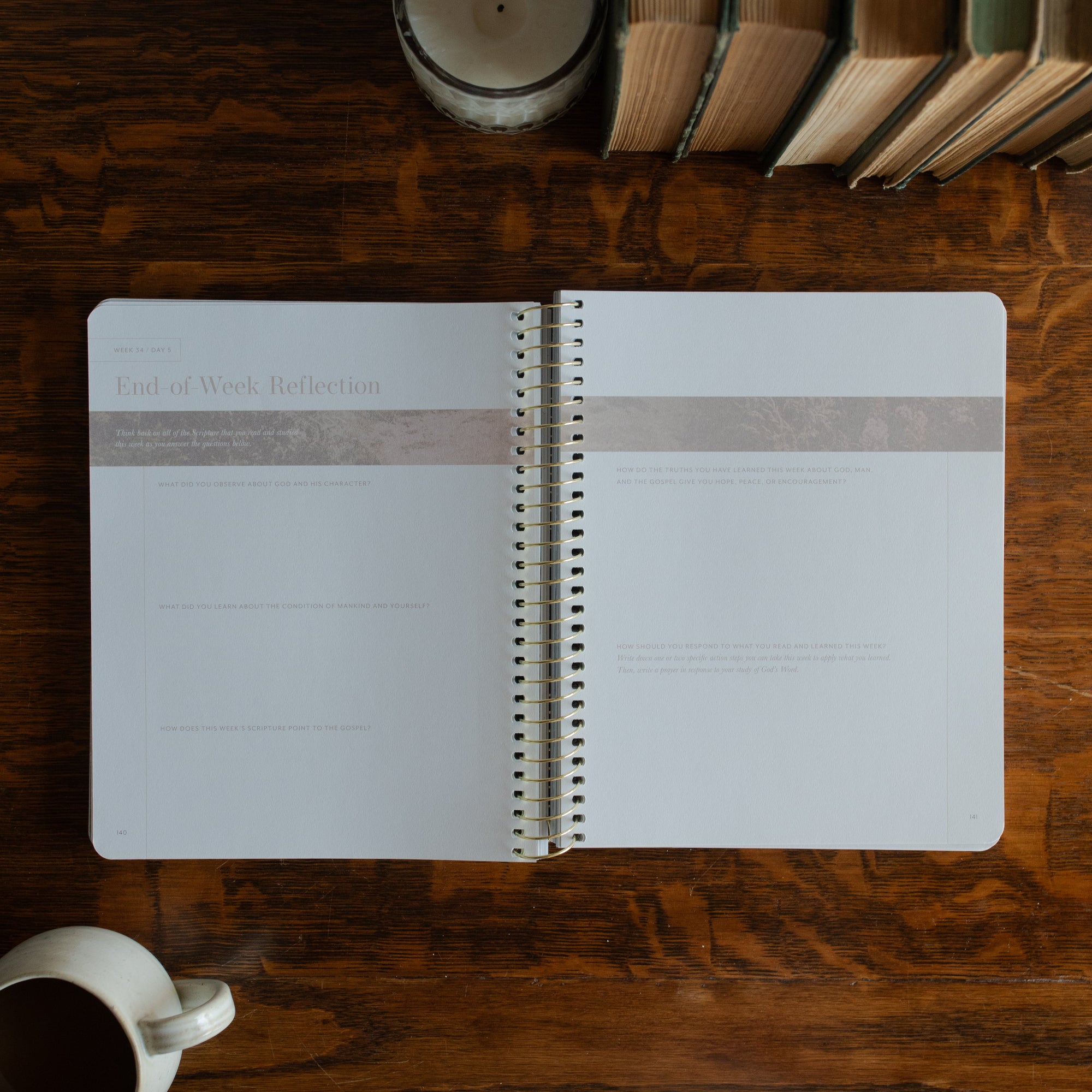 Behold Spiral opened to the end of the week reflection page on a wooden table next to a coffee cup and some books