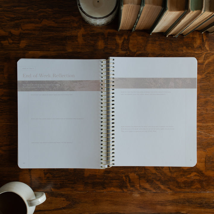 Behold Spiral opened to the end of the week reflection page on a wooden table next to a coffee cup and some books