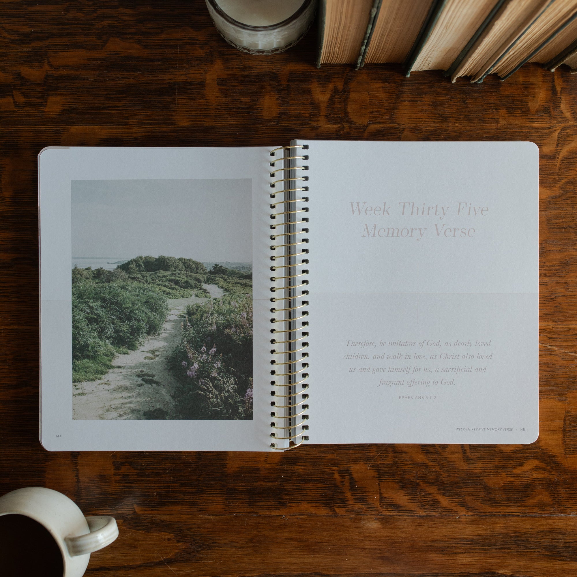 Behold Spiral opened to the memory verse page on a wooden table next to a coffee cup and some books