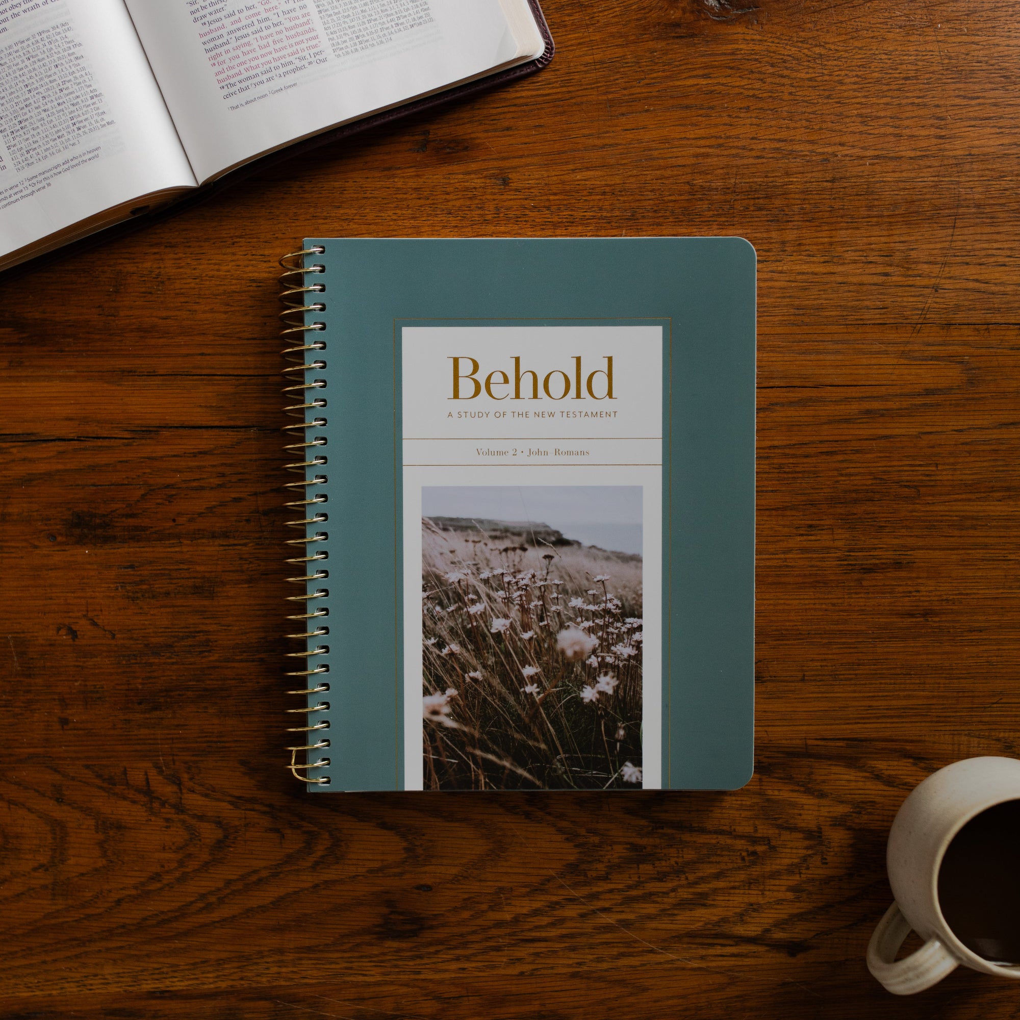 Behold Volume 3 Spiral cover on a wooden table near a coffee cup and an open bible