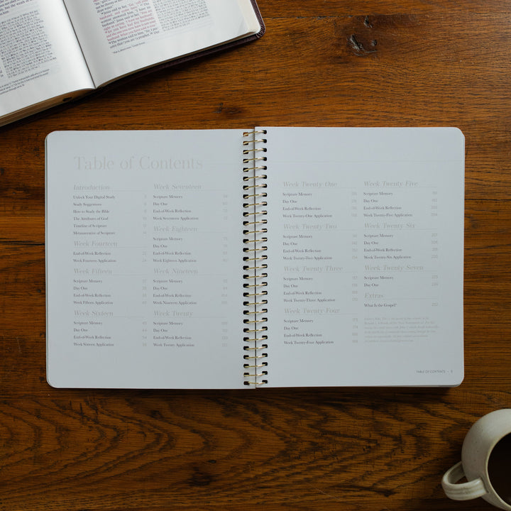 Behold Volume 3 Spiral opened to the table of contents page on a wooden table near a coffee cup and an open bible