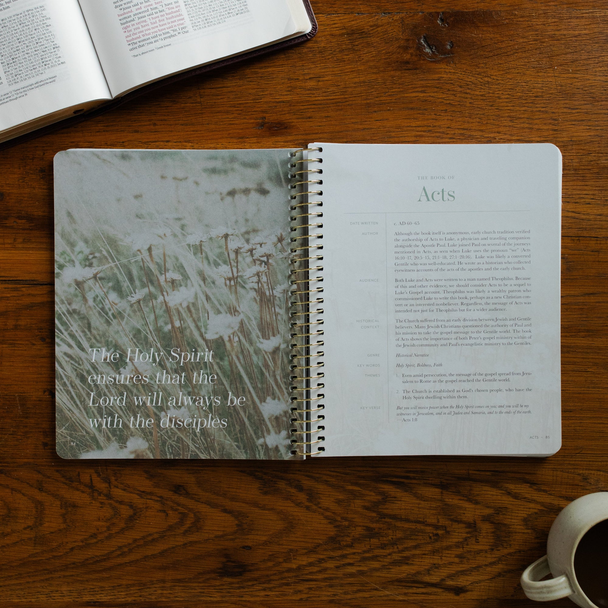 Behold Volume 3 Spiral opened to the book of Acts page on a wooden table near a coffee cup and an open bible