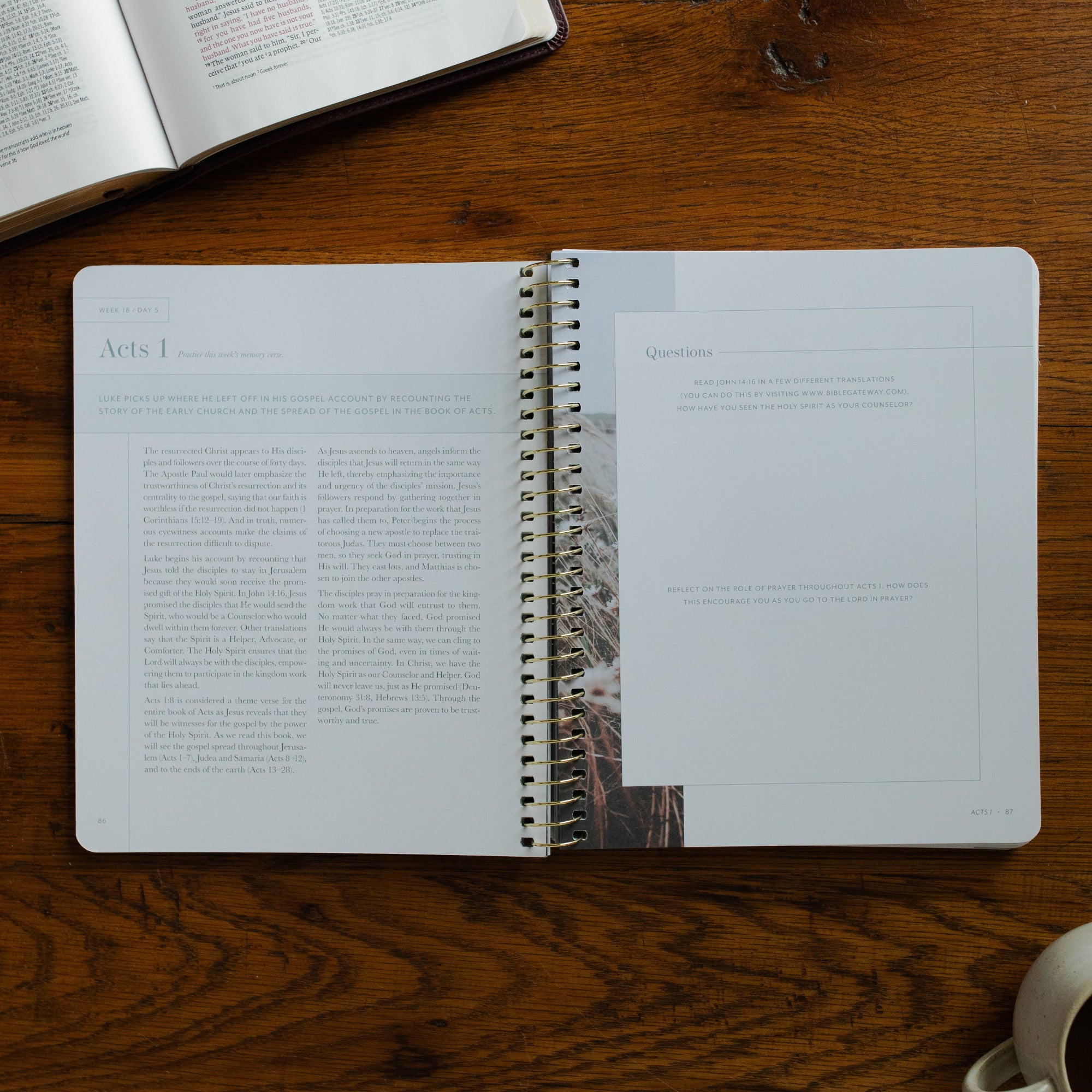 Behold Volume 3 Spiral opened to the questions page on a wooden table near a coffee cup and an open bible