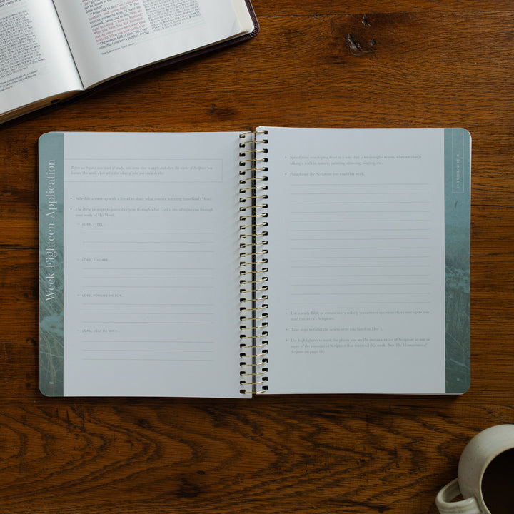 Behold Volume 3 Spiral opened to the applications page on a wooden table near a coffee cup and an open bible