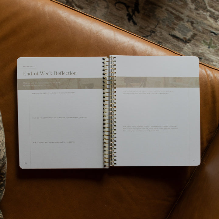 Behold Spiral Study opened to the end of week reflection page on a leather couch