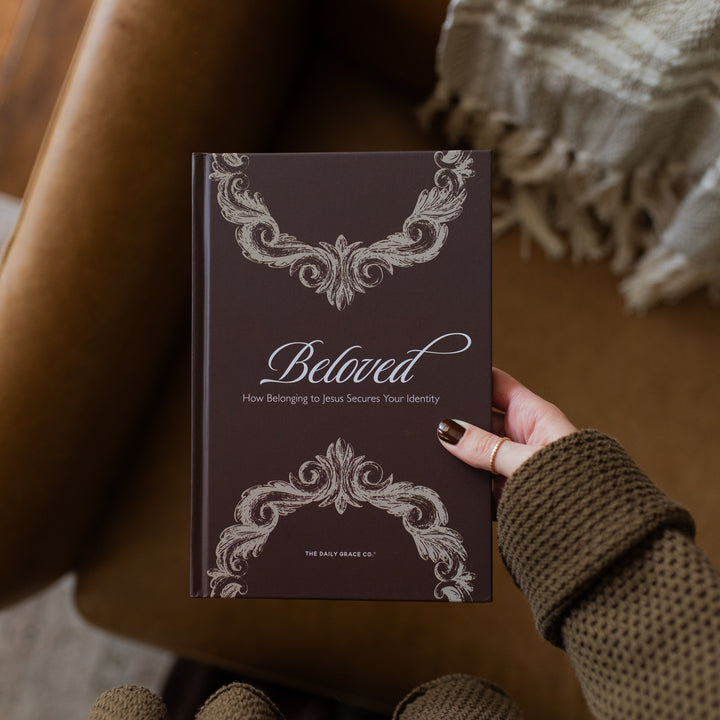 A woman holding the Beloved How Belonging to Jesus Secures your Identity book cover over a brown leather couch