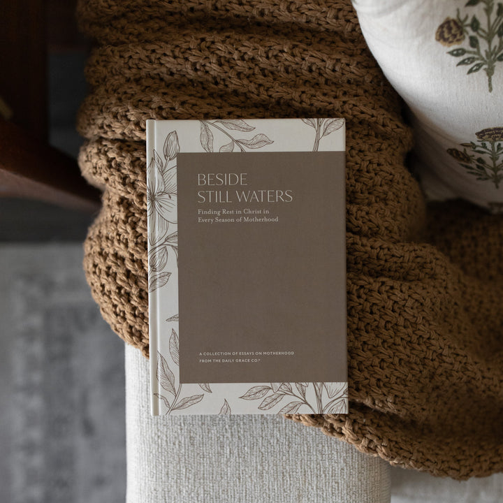 Beside Still Waters Motherhood Book Cover on a brown knitted blanket on the arm of a white couch