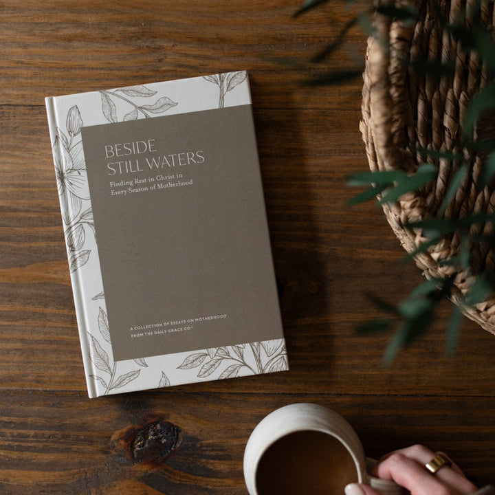 Beside Still Waters Motherhood Book Cover on a brown wooden table next to a white coffee cup being held by a woman's hand and a woven basket