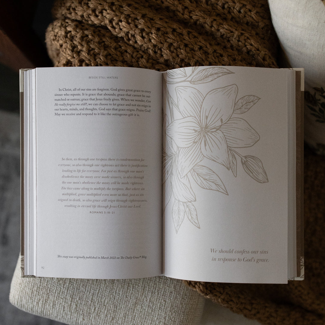 Beside Still Waters Motherhood Book opened to a page 92, with a floral design on the other page, on a brown knitted blanket on the arm of a white couch