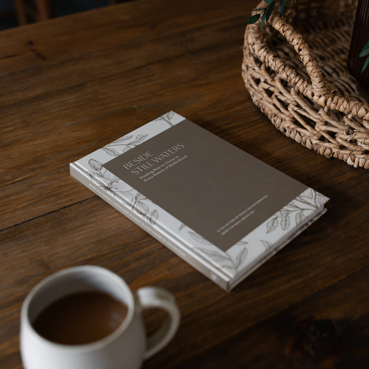 Beside Still Waters Motherhood Book Cover on a brown wooden table next to a white coffee cup and a woven basket