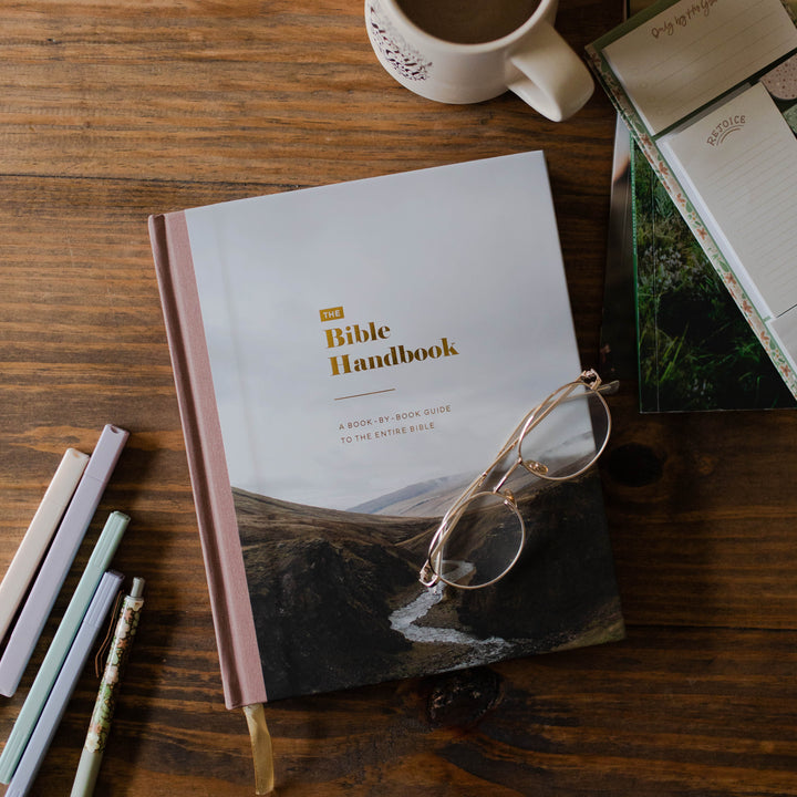 A Bible Handbook notebook with a pinkish-brown spine and a landscape cover, styled on a wooden desk with a coffee cup, glasses, and pens.