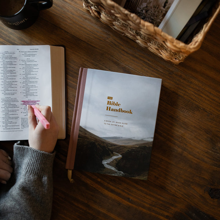 A Bible Handbook book lies on a dark wooden table next to an open Bible, with a hand holding a pink highlighter. A black mug and a woven basket are also visible in the scene.