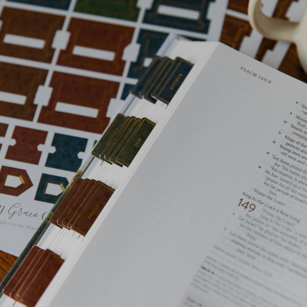 An open Bible with printed pages visible and colored divider tabs arranged beside it on a patterned surface. The tabs appear in dark blue, green, and brown tones, and the surrounding area includes a multicolored geometric mat or book cover.