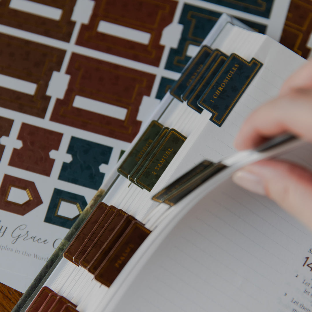 A close-up of a hand organizing printed index cards or a planner with brown, teal, and gold tabbed sheets over a white lined page.