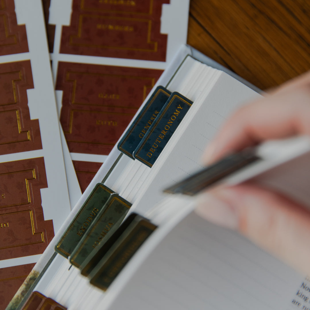 A close-up of a hand placing a dark blue index tab labeled 'PETERMONOM' into a lined notebook. Other matching index tabs in gold and green are visible, along with sheets of the tabs on a wooden table.