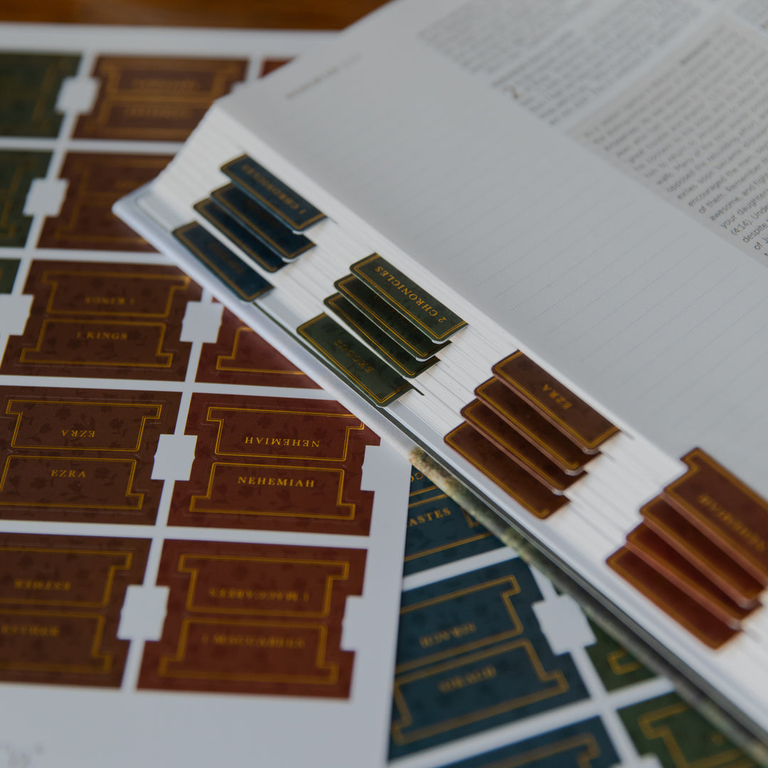 A close-up of a book or album page with decorative gold and brown library-style tabs, placed over printed sheets showing matching tab designs.