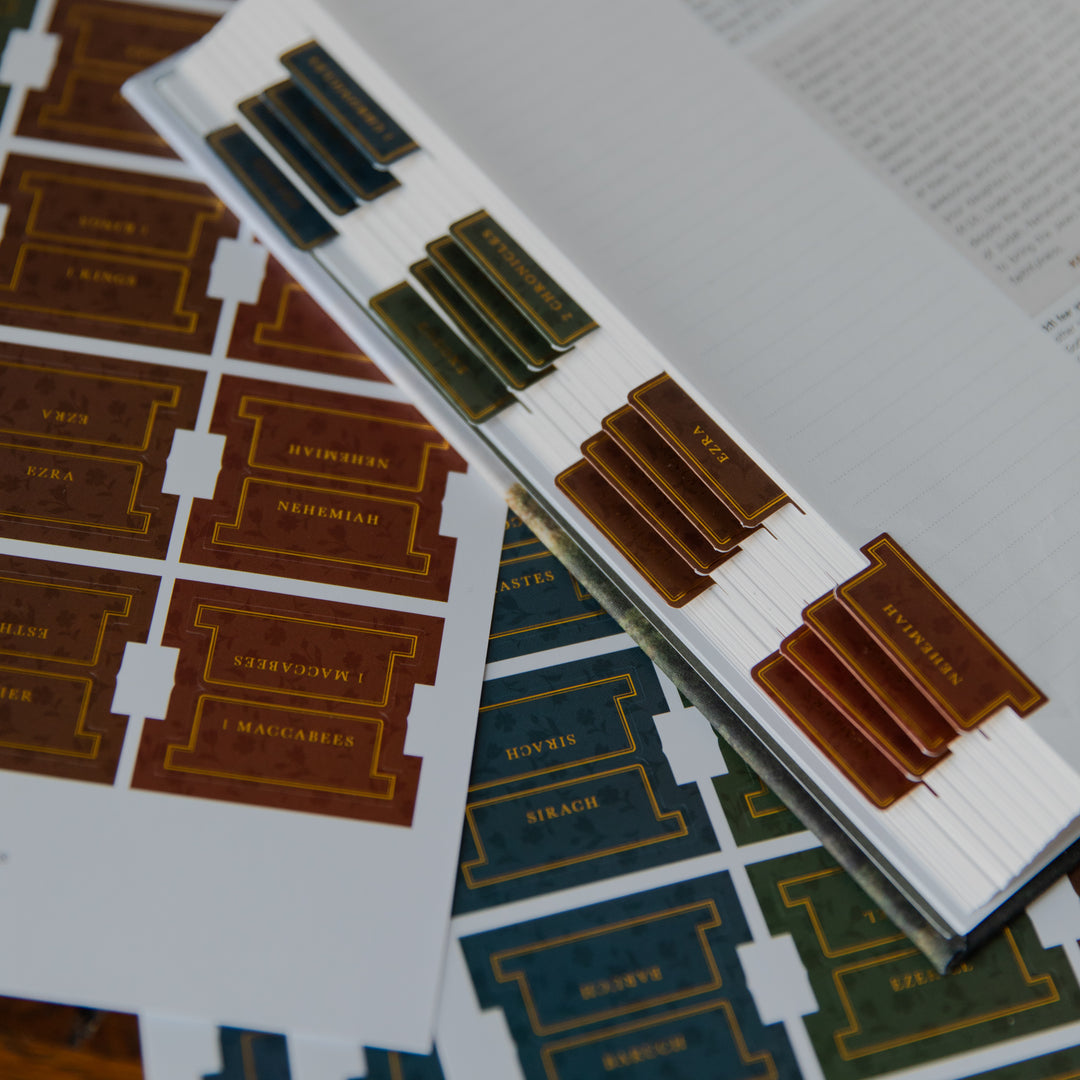 A close-up of printed Hebrew nameplates or labels arranged on sheets, with some labels placed on top of an open book. The visible labels are rectangular with gold decorative borders and text, shown in brown, dark green, and blue tones against white sheets.