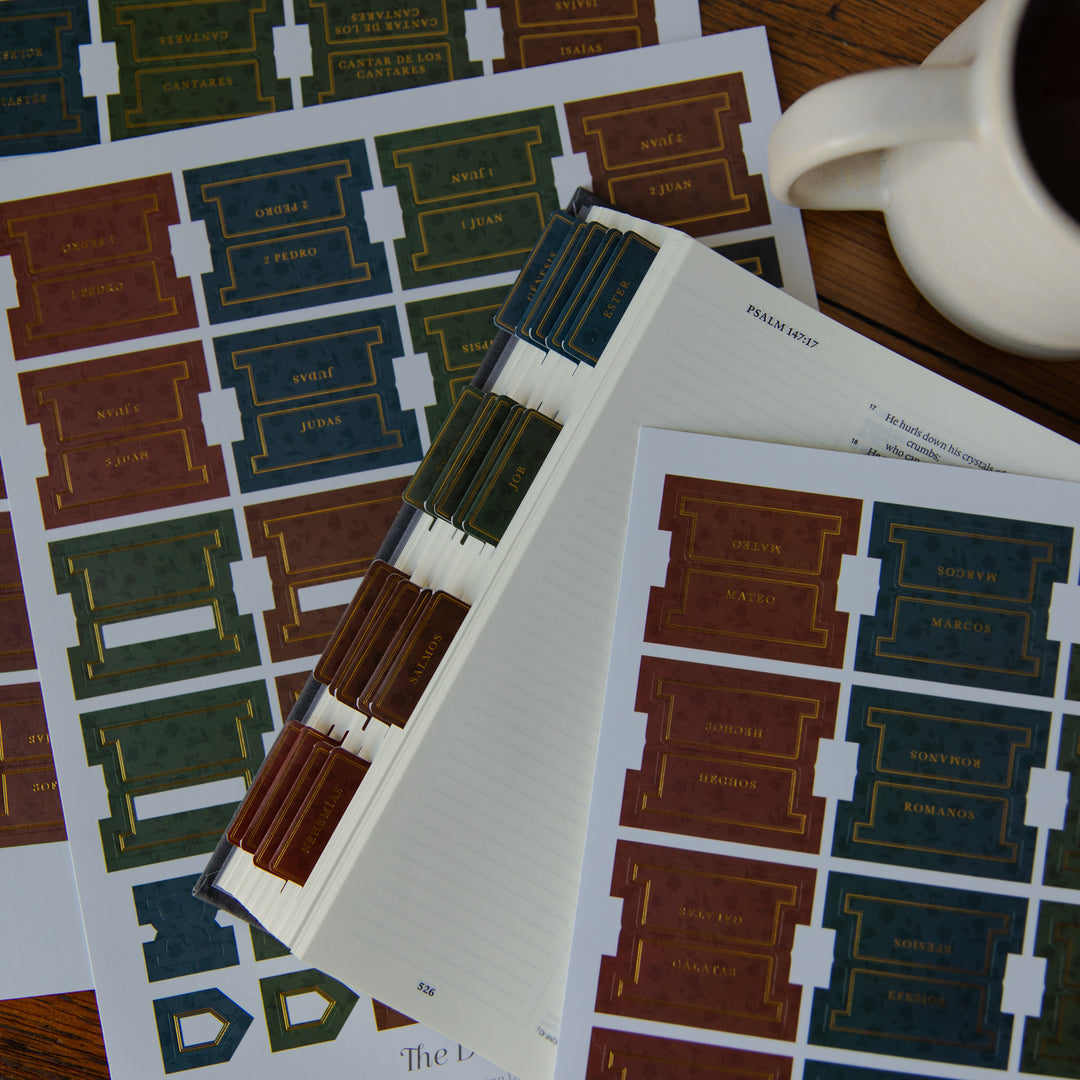A styled flat-lay of a bookbinding workshop or craft product, featuring sheets of colorful paper labels and a small stack of bound books. A white ceramic mug is visible in the top right corner on a wooden surface.