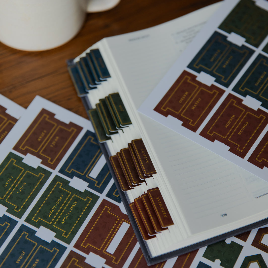 A close-up of an open notebook or planner showing printed tab inserts for organizing pages, with matching printed sheet designs spread on a wooden surface. A white mug is partially visible at the top left.