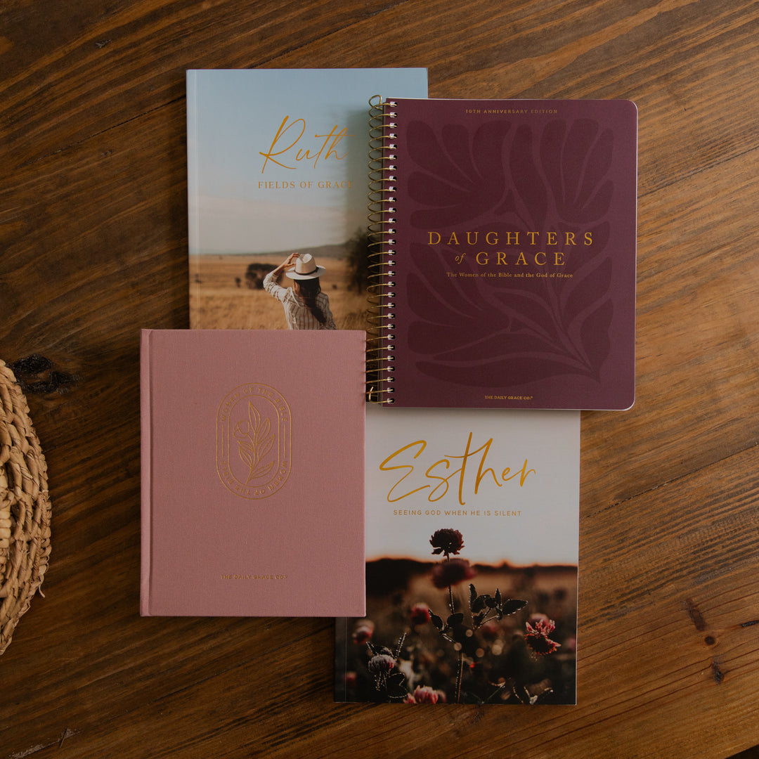 Four inspirational journals from the brand 'The Devoted Wife' arranged on a rustic wooden table. The collection includes three spiral-bound notebooks in dusty rose, burgundy, and light blue, alongside one hardcover journal in dusty rose with gold foil detailing.