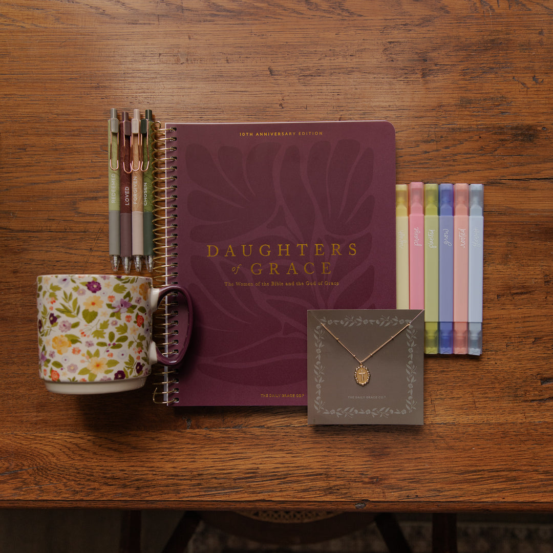 A styled flat-lay on a wooden table featuring a maroon spiral-bound journal with gold lettering that reads 'Daughters of Grace' and '150th Anniversary Edition.' Surrounding it are a floral ceramic mug, a set of metallic pens, pastel highlighters, and a small jewelry card holding a gold pendant necklace.