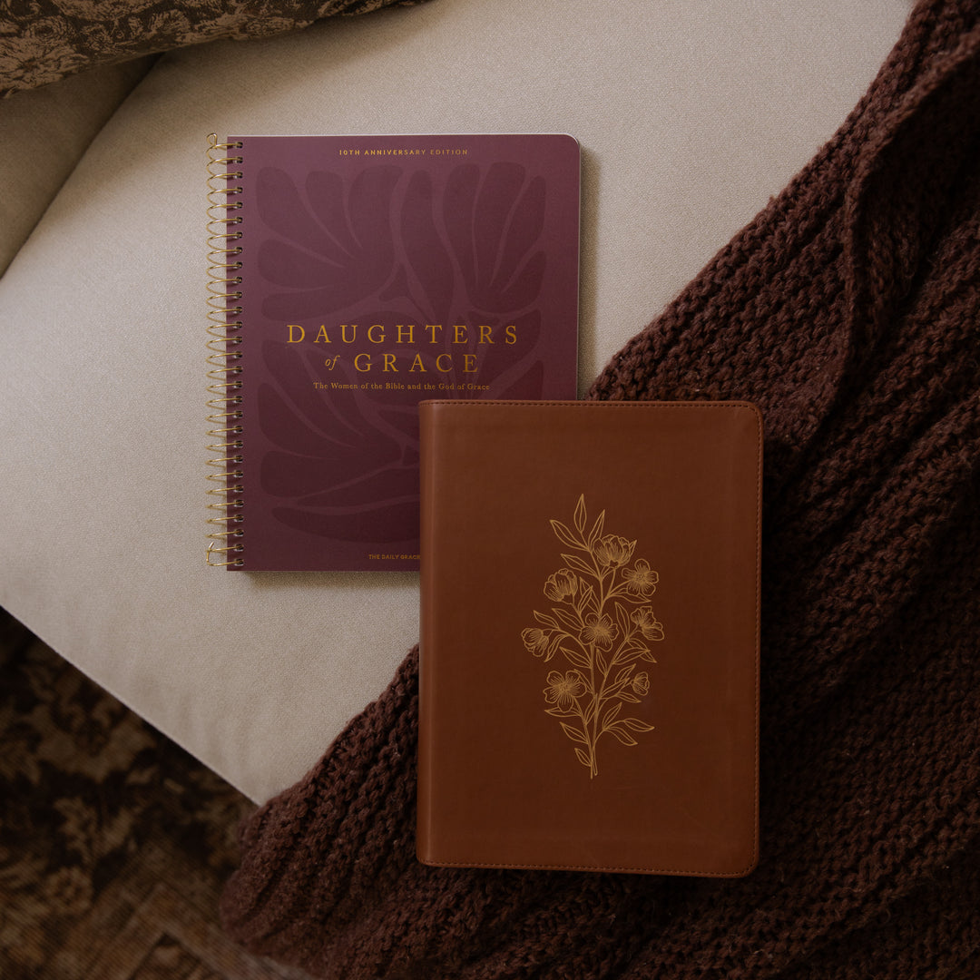 A journal and a bible styled on a neutral fabric surface with a chunky knit brown throw blanket. The back cover of one journal shows the title 'DAUGHTERS OF GRACE' in gold lettering, and the front cover of the other is brown with a gold floral engraving.