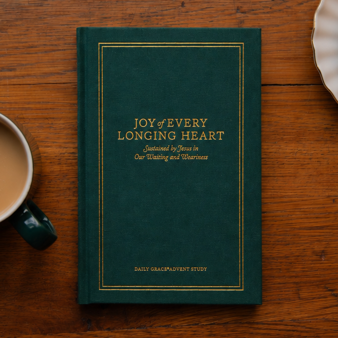 A dark green hardcover book titled 'Joy of Every Longing Heart' lies on a rustic wooden table, styled with a cup of coffee, a chocolate chip cookie on a plate, and green pine branches at the top.