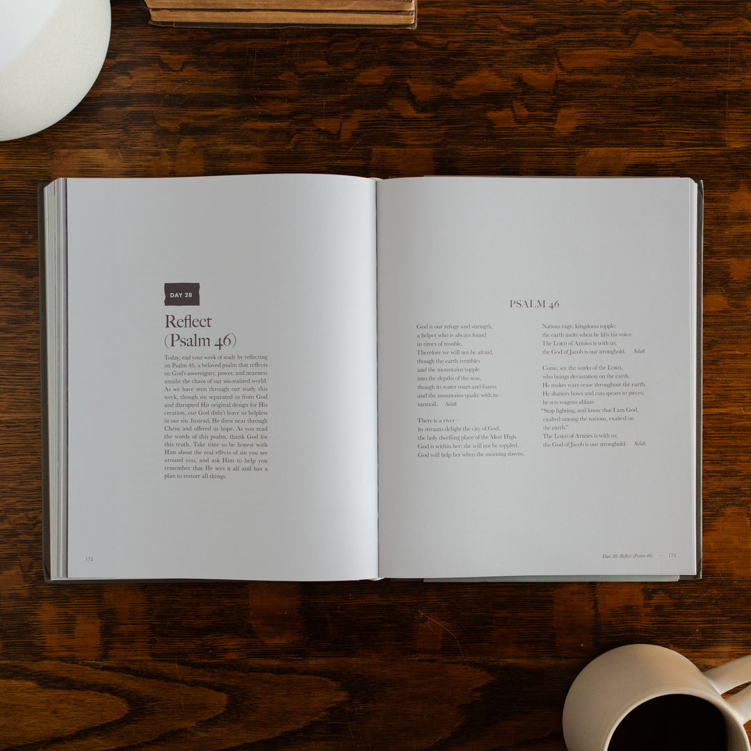 Clinging to the Cross Spiral hardcover opened to the Reflect(Psalm 46) page on a wooden table next to a coffee cup