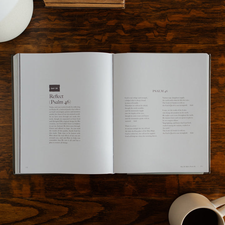 Clinging to the Cross Spiral hardcover opened to the Reflect(Psalm 46) page on a wooden table next to a coffee cup