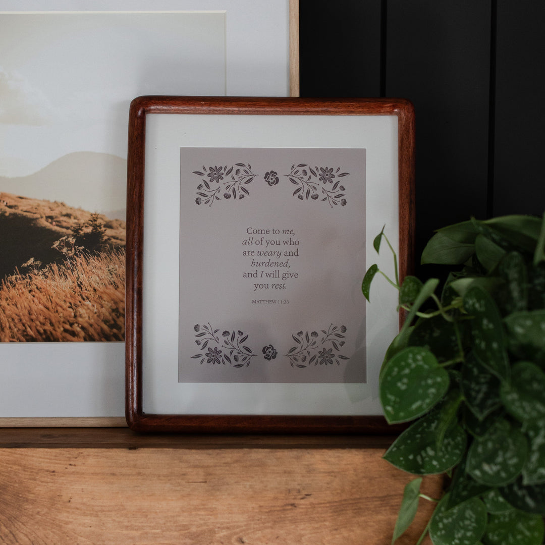 Come to Me Floral Print in a brown wooden frame leaning on another photo; The Print is siting upright on a wooden table next to some greenery