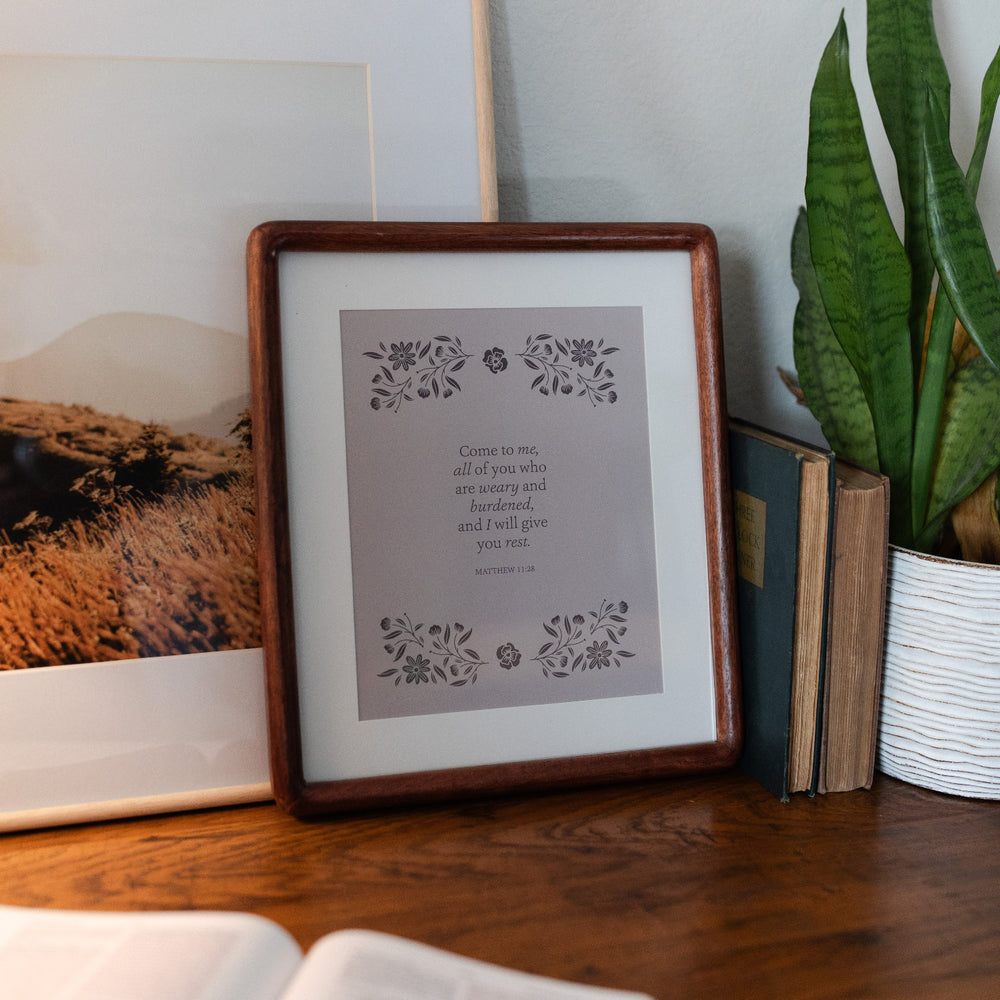 Come to Me Floral Print leaning against another photo in a frame; Two vintage books and a plant in a white vase are next to the floral print with an open bible at the foot of the print, all resting on a wooden table