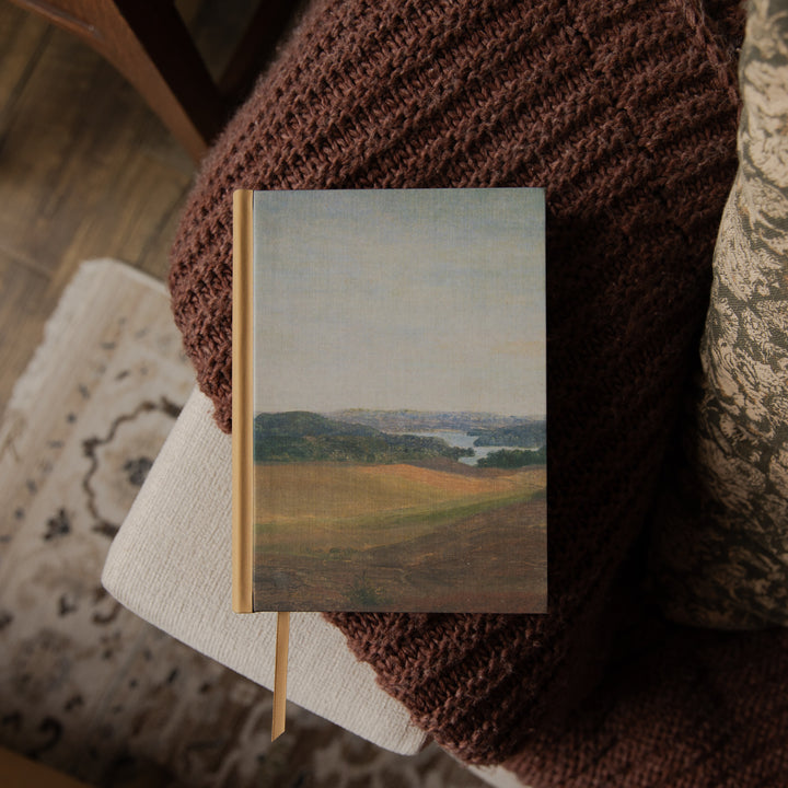 A hardcover bible with a landscape painting cover, resting on a cozy brown knitted blanket over a light-colored cushion.