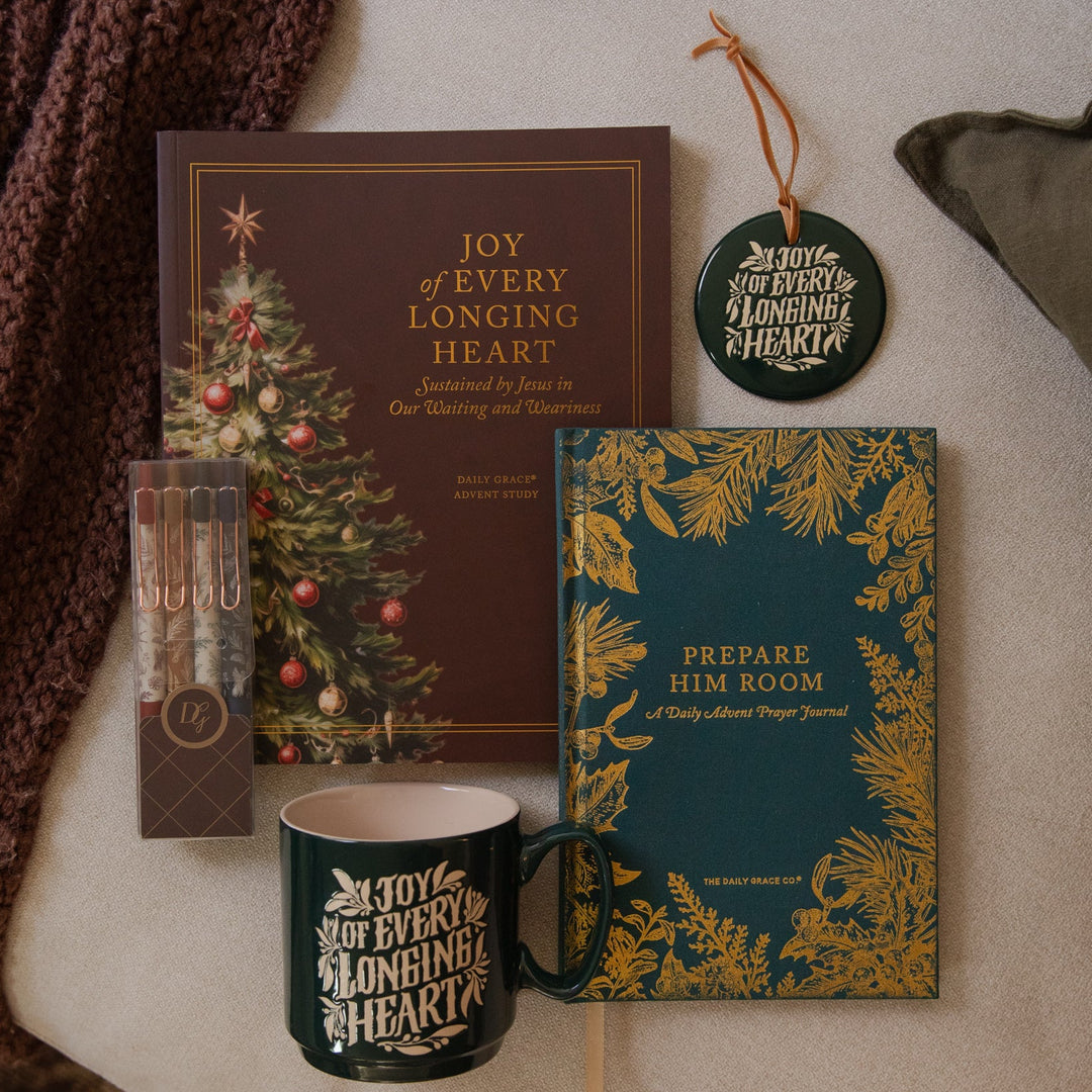 A styled flat-lay of coordinated holiday stationery and home decor items on a light fabric surface, including a brown book with a Christmas tree illustration, a teal journal with gold botanical print and the phrase 'Prepare Him Room', a dark green mug with white holiday lettering, a small boxed set of pens or markers, a round hanging ornament, and a chunky knit brown blanket.